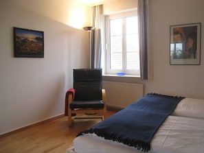 A room with a bed, a chair, a window with curtains, and two framed pictures on the walls.