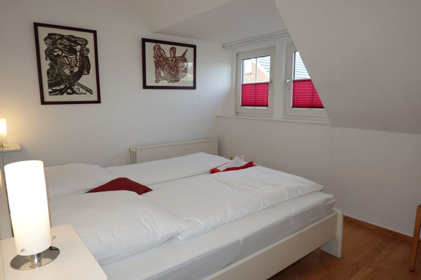 Ein Schlafzimmer mit einem Bett, zwei gerahmten Kunstwerken, einem Fenster mit roten Jalousien und einer Nachttischlampe.