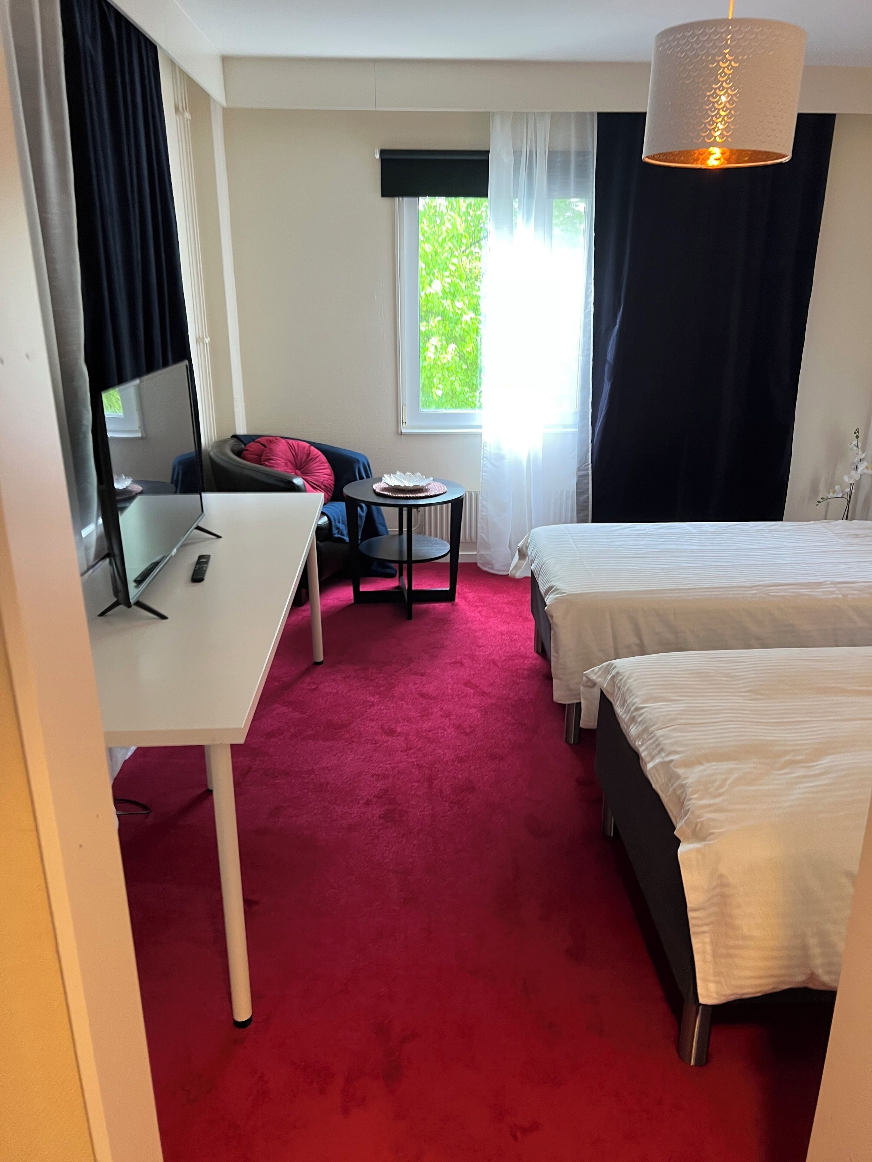 A hotel room with a desk, two beds, a small table, and a window with curtains.