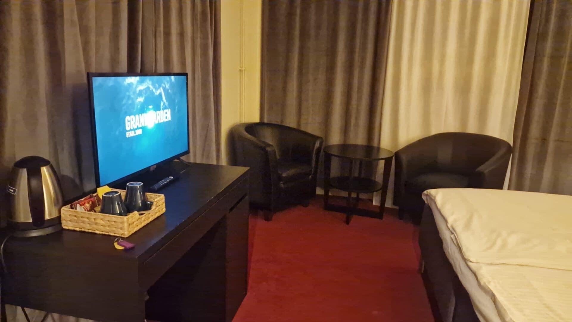 A hotel room with a TV displaying 'GRAND GARDEN ETAL 2006', a bed, two chairs, a small table, and a coffee maker.