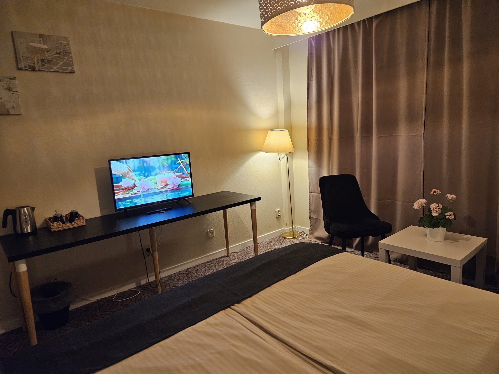 A hotel room with a bed, a desk with a television, a chair, a small table with a vase of flowers, and a lamp.