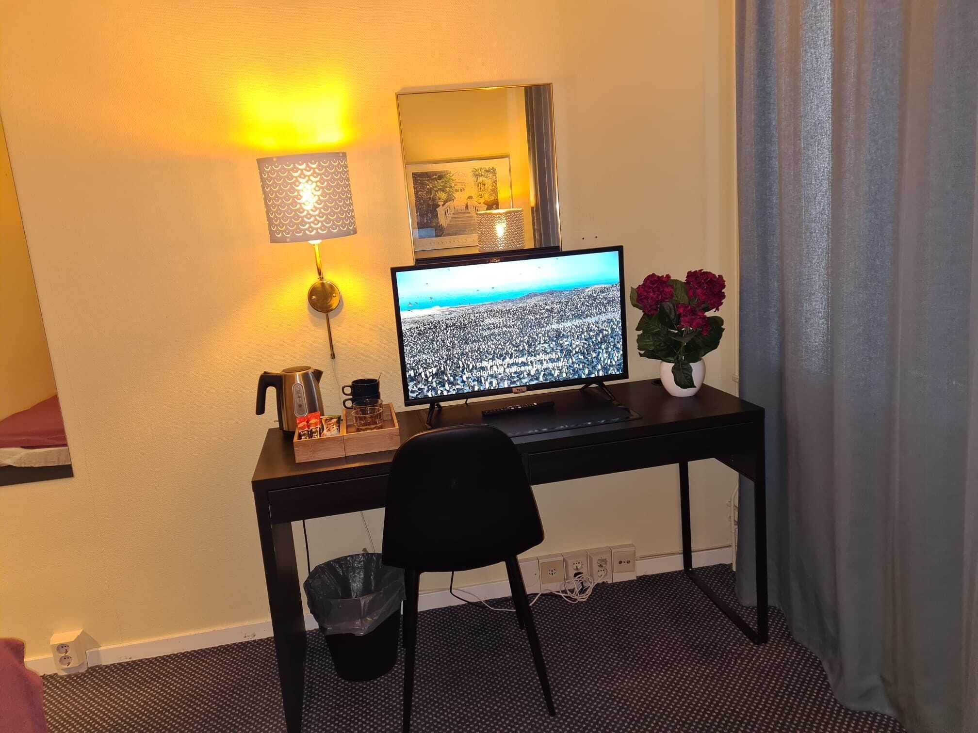 Habitación de hotel con escritorio, monitor de computadora, cafetera, cubo de basura y un florero con flores.