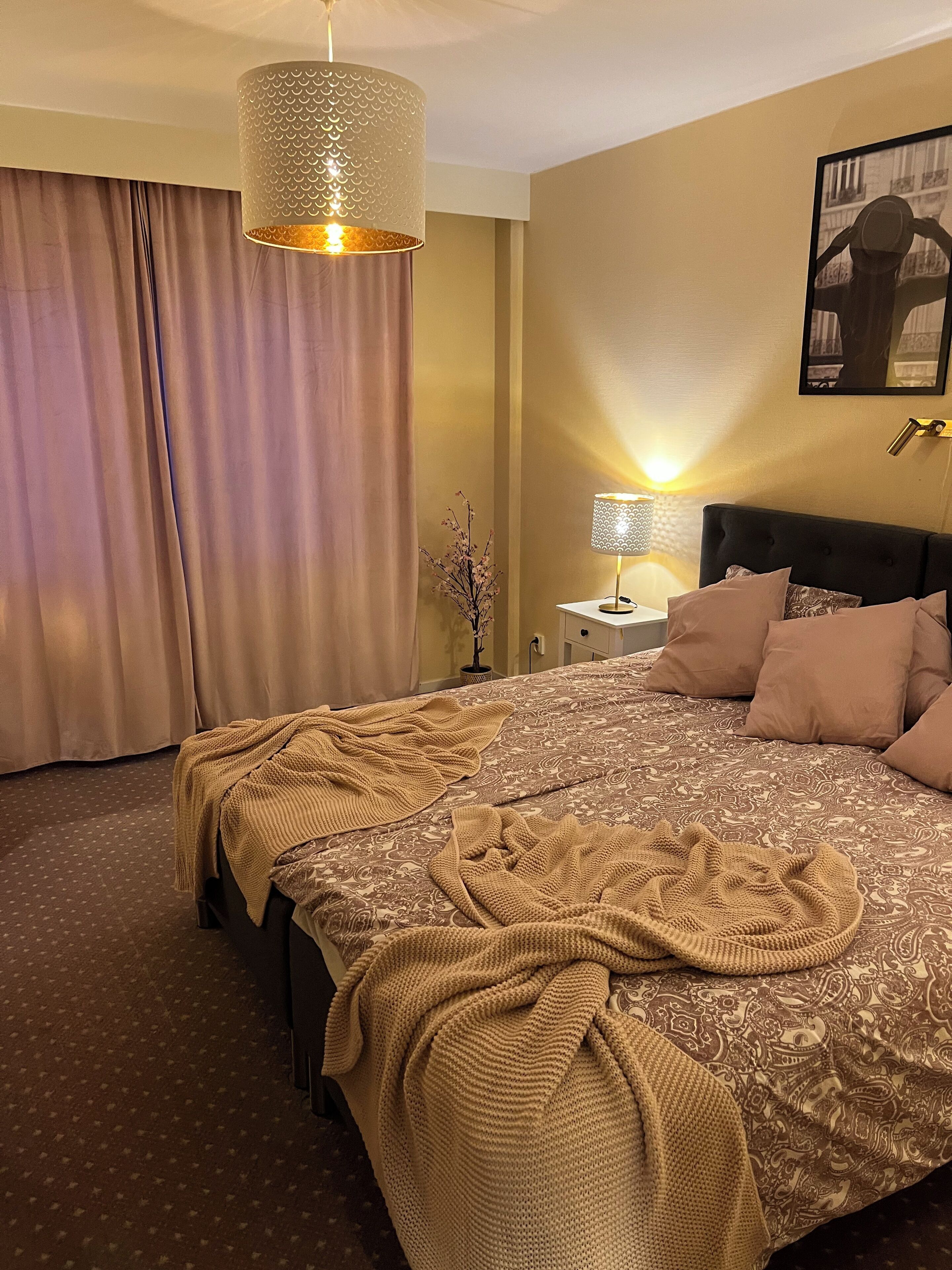 Ein Hotelzimmer mit einem großen Bett, einem Nachttisch mit Lampe, einer Hängeleuchte und einem gerahmten Bild an der Wand.