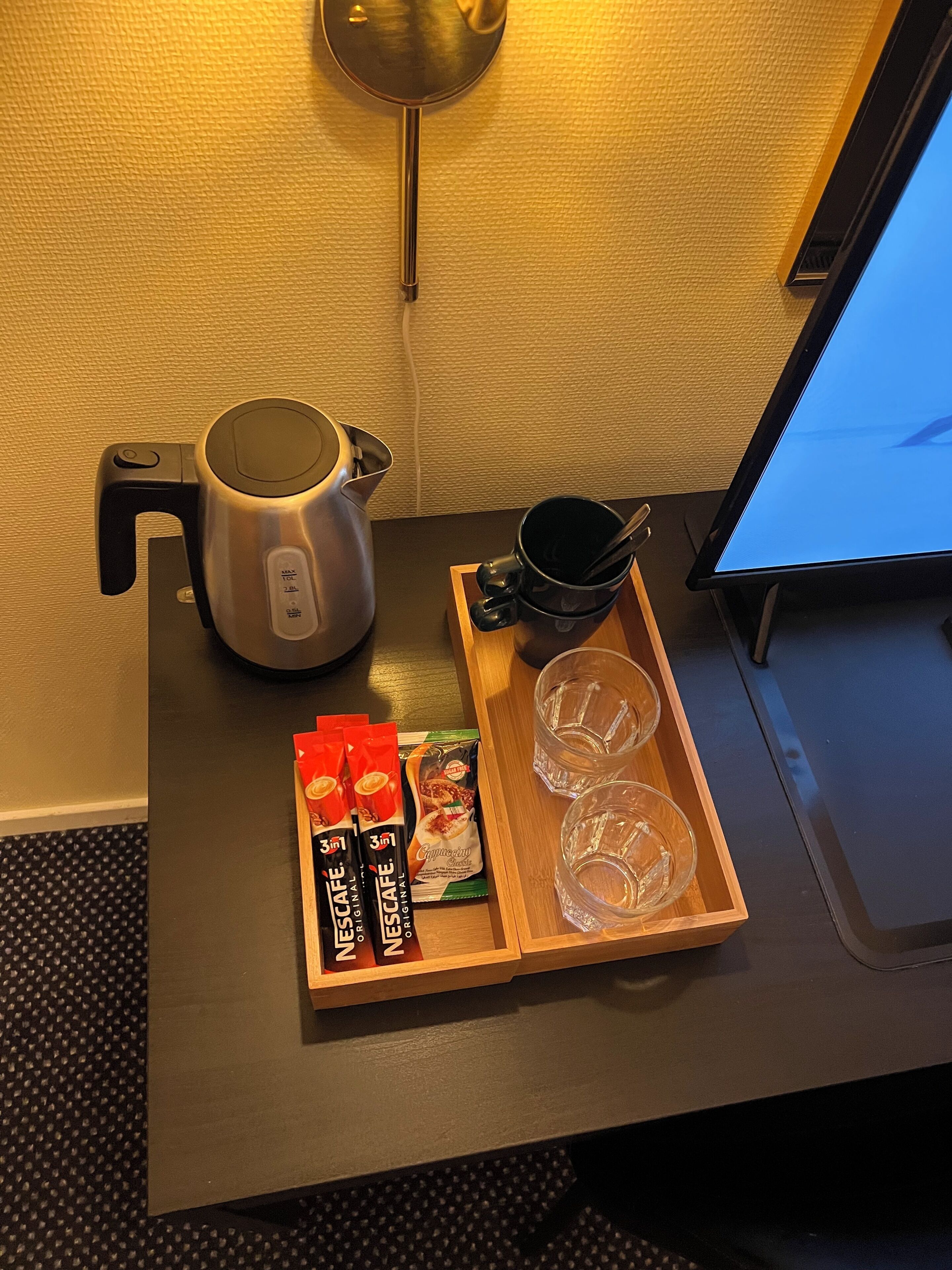A hotel room with a wooden tray containing Nescafe capsules, a black mug, and two clear glasses.