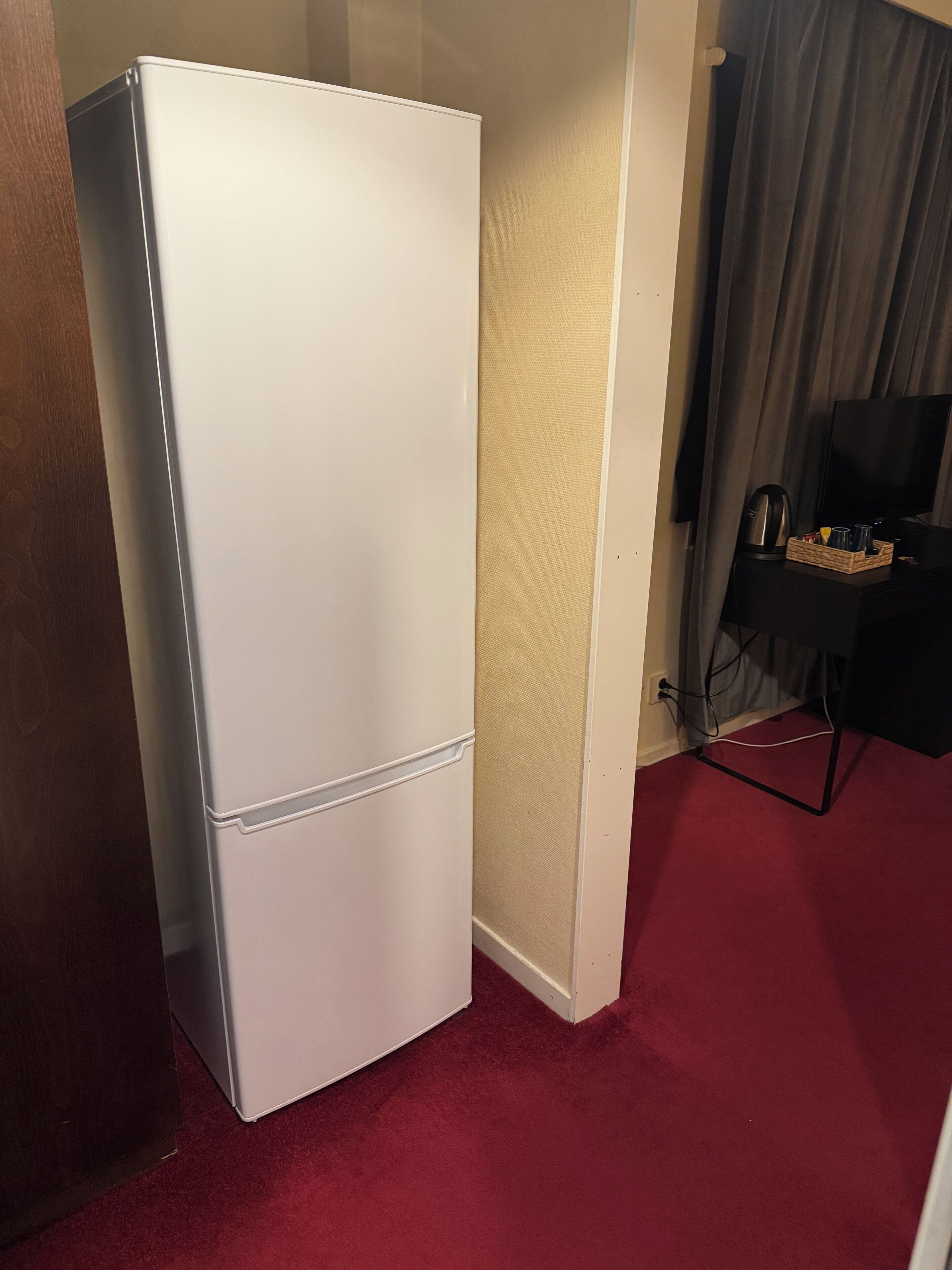 Ein weißer Kühlschrank in einem Raum mit rotem Teppich, einem Fernseher und einem Tablett mit Tassen und einem Wasserkocher auf einem Tisch.