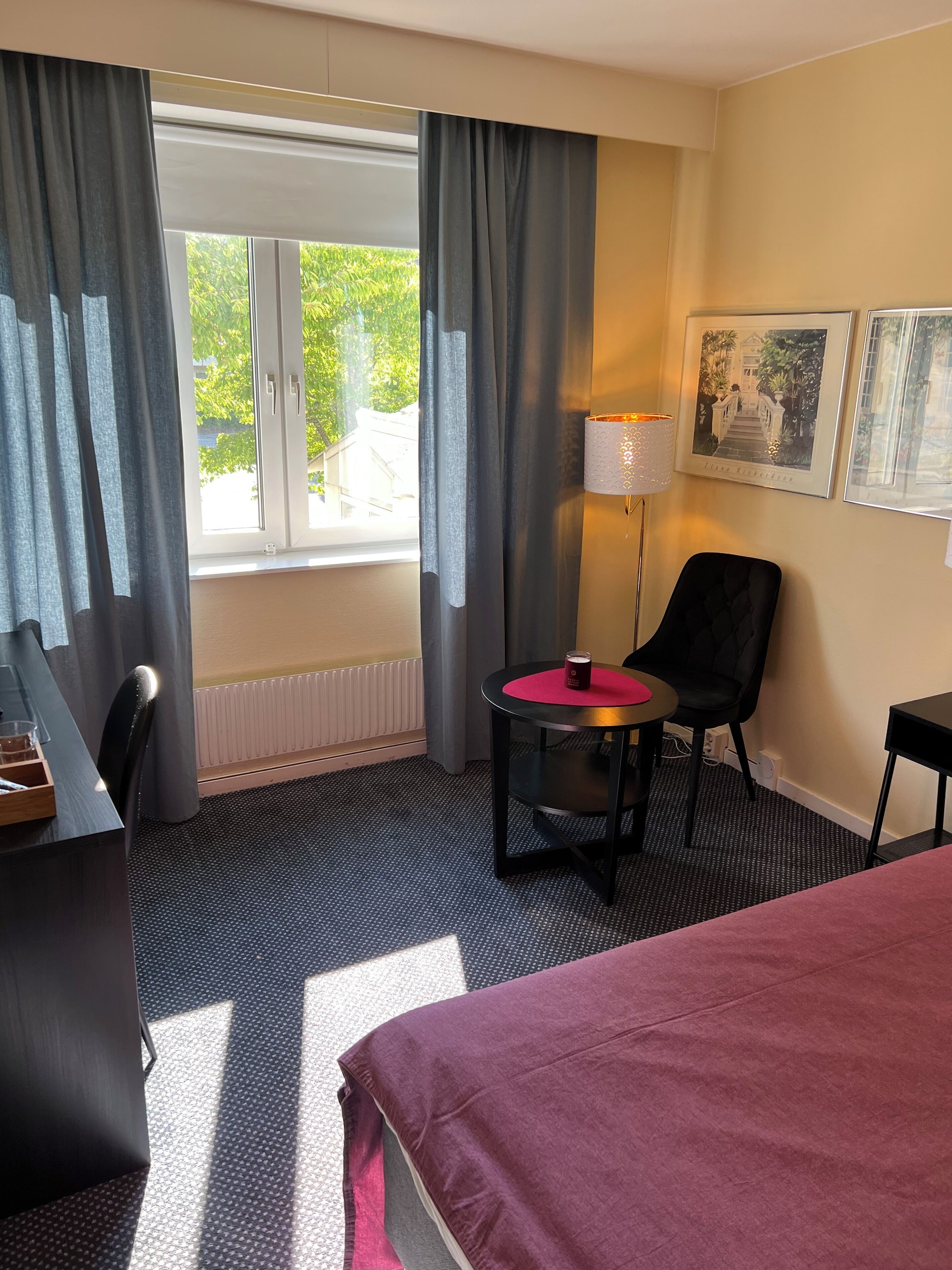 Hotellihuone, jossa on sänky, työpöytä, tuoli, pieni lamppupöytä ja ikkuna, jossa on verhot.