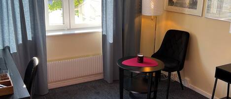 A hotel room with a bed, a desk, a chair, a small table with a lamp, and a window with curtains.
