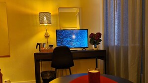 A hotel room with a desk, a computer, a chair, a table with a red coaster, a lamp, and a vase with flowers. - Noah Longstay  (Vasteras)