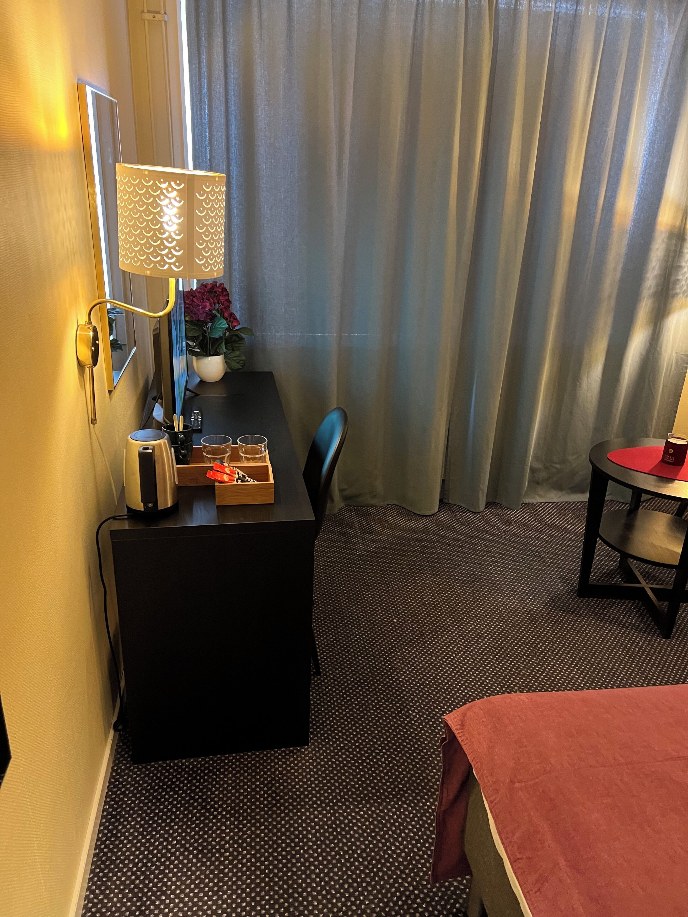 Habitación de hotel con escritorio, lámpara, silla, mesita con mantel rojo y cama con manta roja.