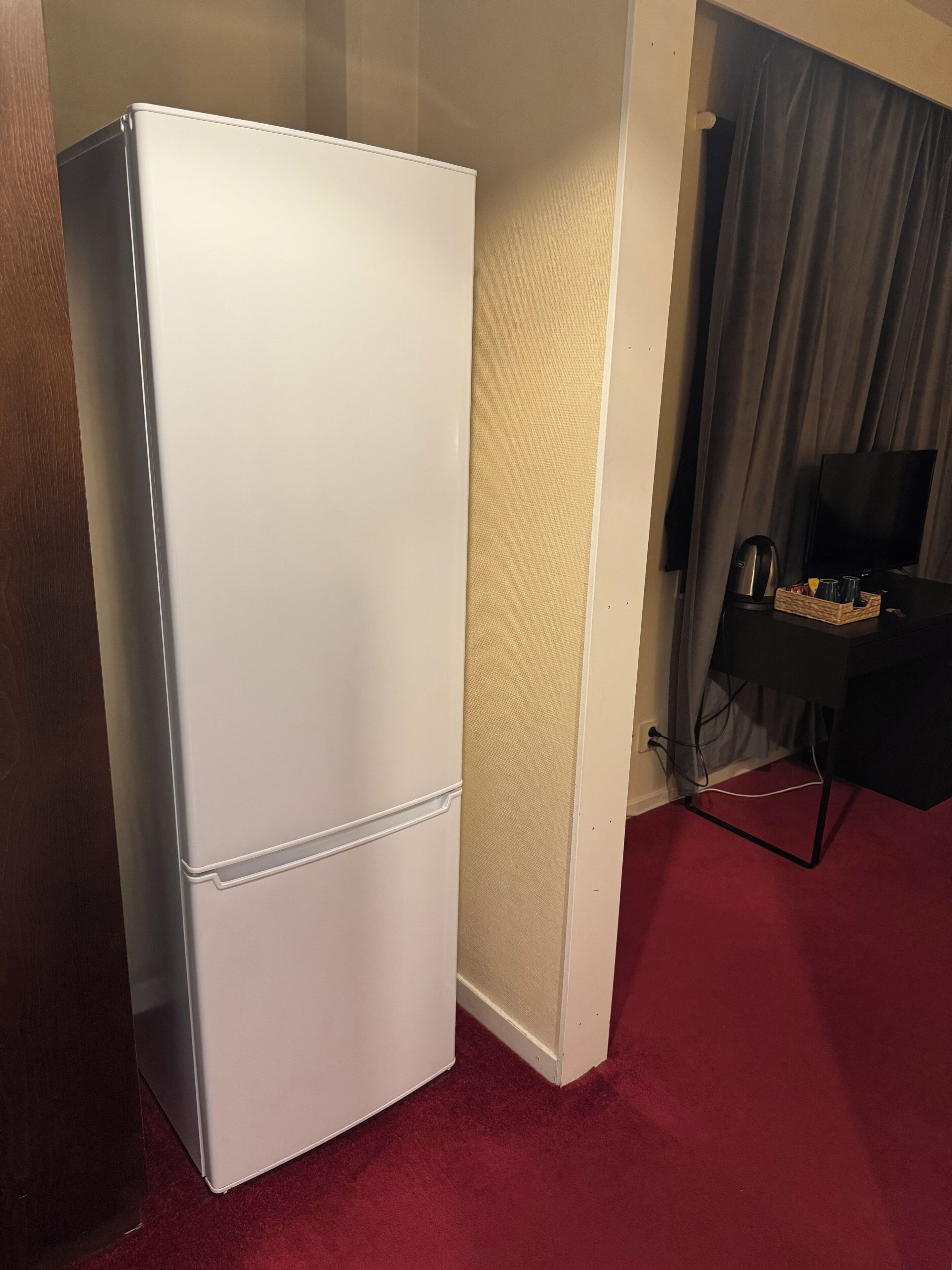 A white refrigerator in a room with a red carpet, a television, and a small table with a kettle and cups.