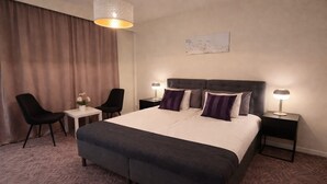 A hotel room with a large bed, two bedside tables with lamps, a desk with a chair, and a large window with curtains. - Noah Longstay  (Vasteras)