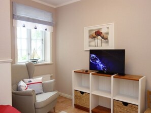 Un salon avec un canapé gris, un téléviseur posé sur un meuble en bois, un tableau encadré accroché au mur et une fenêtre donnant sur un paysage verdoyant.