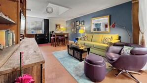 Un salon avec un canapé, une table basse et des fauteuils violets. On y trouve également une table à manger avec des tabourets, une bibliothèque et un téléviseur.