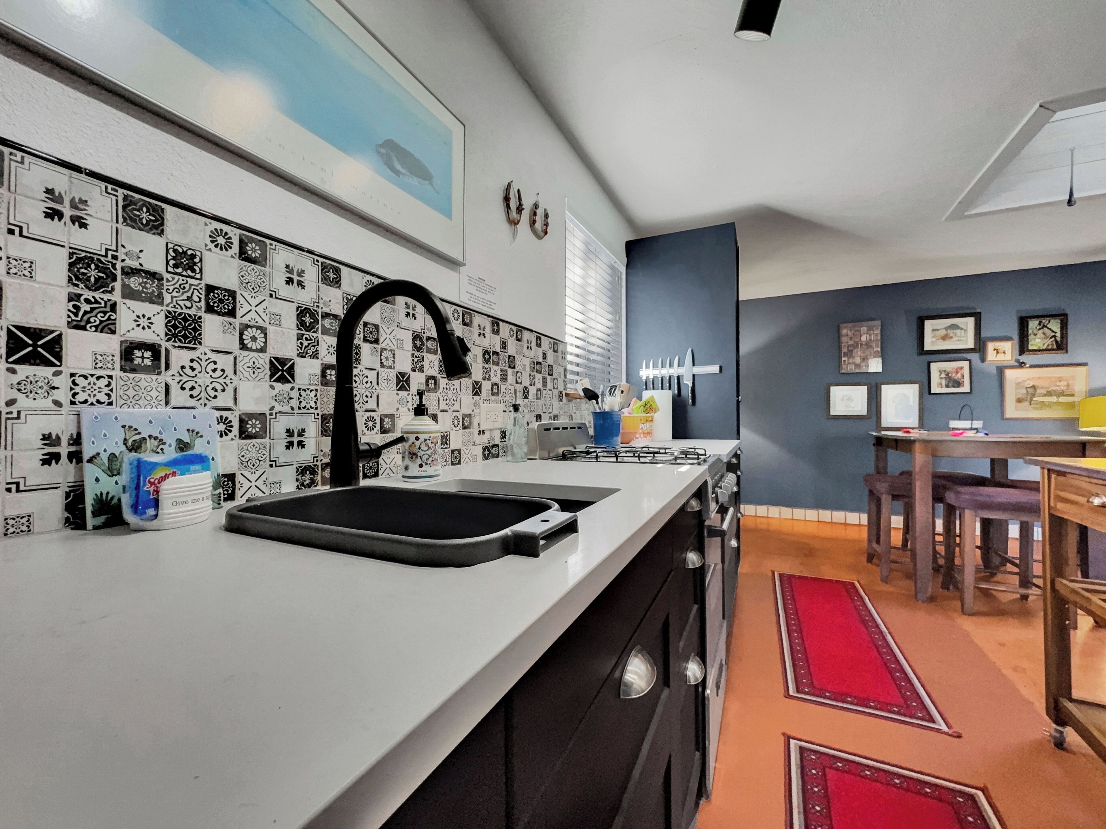 Een keuken met een wit aanrecht, een zwarte kraan en een decoratieve spatwand. Er is een eethoek met een tafel en stoelen, en een rood vloerkleed.