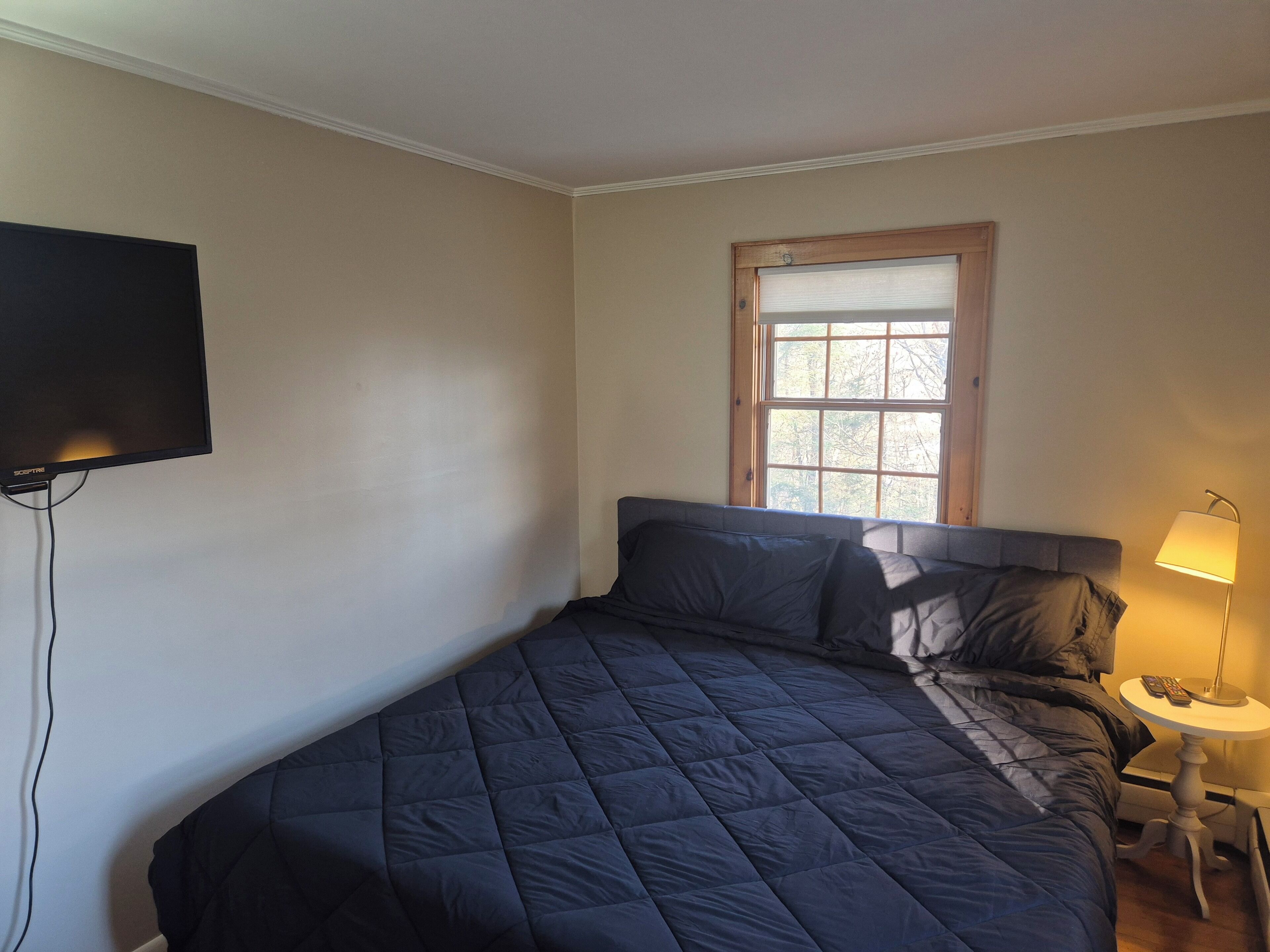 A bedroom with a bed, a television mounted on the wall, a window, and a bedside table with a lamp.