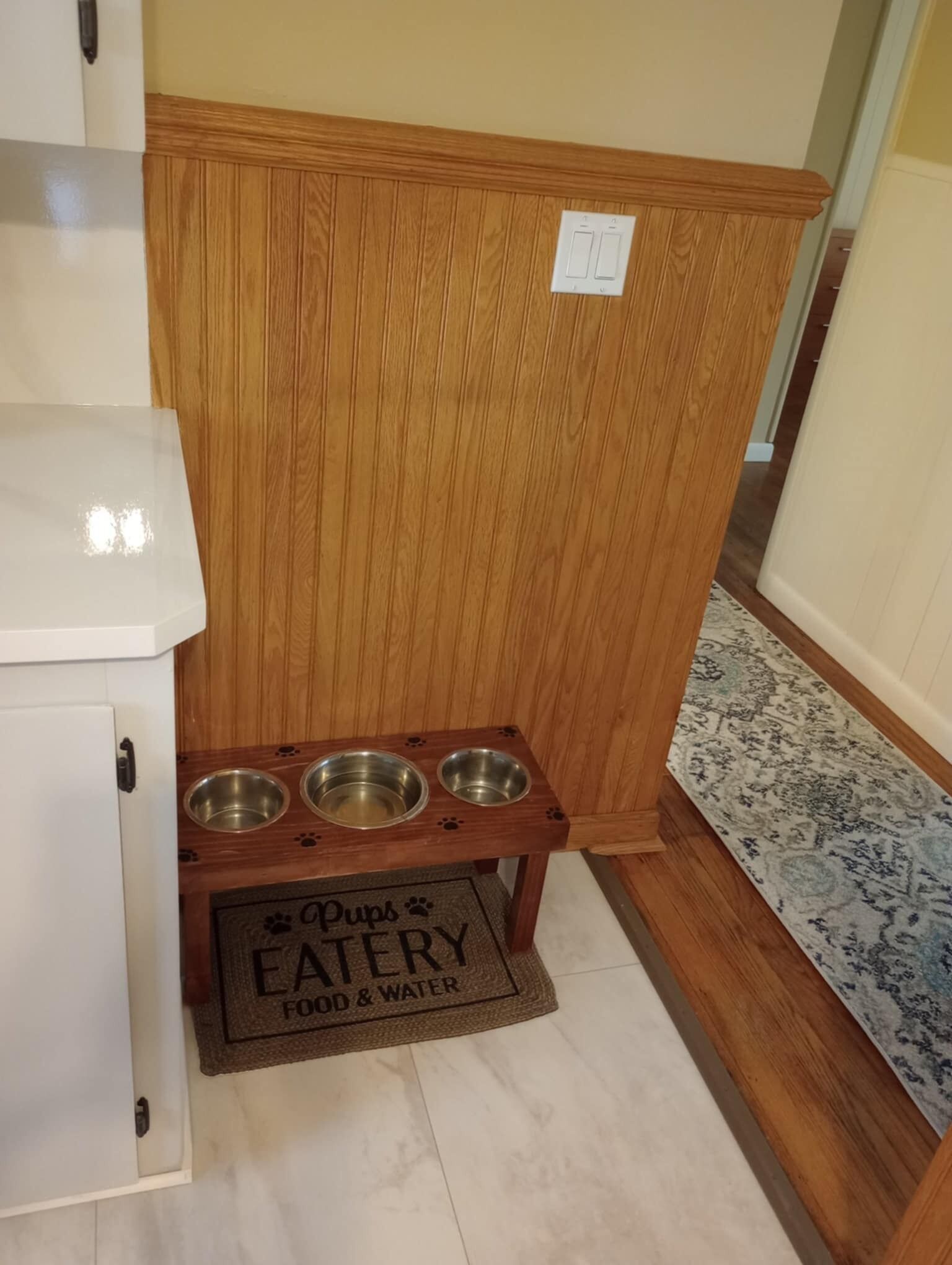A wooden cabinet with three metal bowls on a matching tray, a doormat with "Pups Eatery Food & Water" text, and a patterned rug on the floor.
