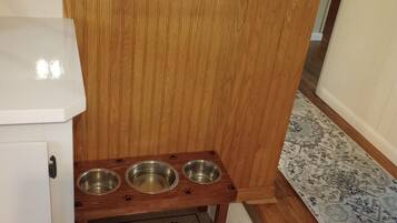 A wooden cabinet with three metal bowls on a matching tray, a doormat with "Pups Eatery Food & Water" text, and a patterned rug on the floor.