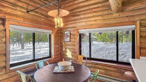 Interior de una cabaña de madera con una mesa redonda, sillas verdes y ventanas amplias que ofrecen vistas de un paisaje nevado.