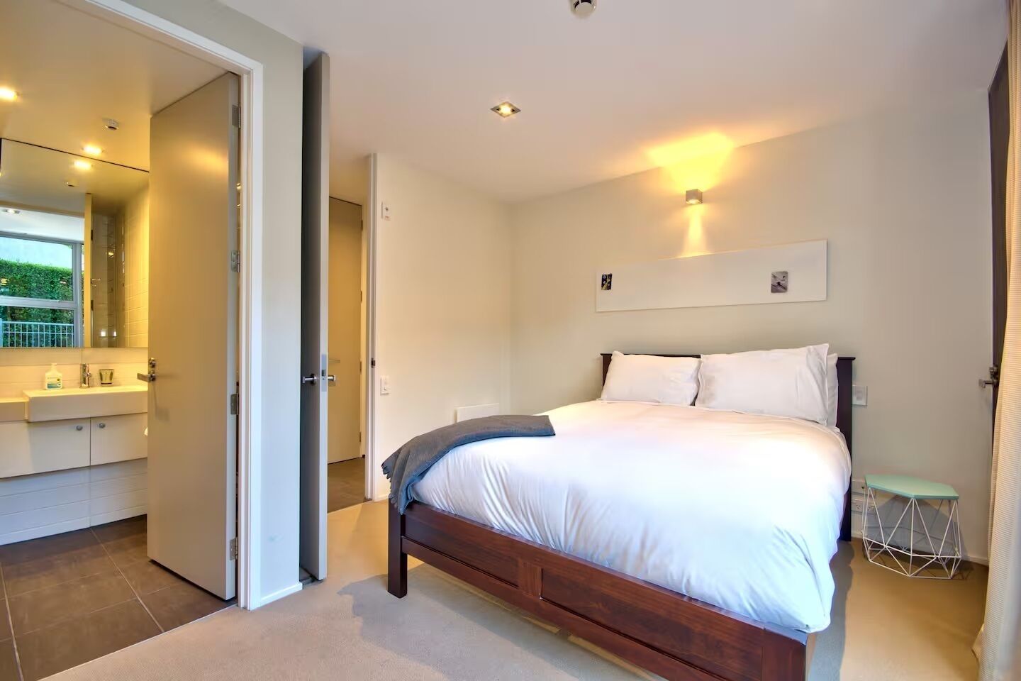 A bedroom with a large bed, a bedside table, a nightstand, a wall-mounted light, and a door leading to a bathroom.