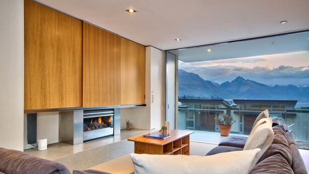 A modern living room with a fireplace, a sofa, and a view of mountains through large windows. - Central Queenstown • 5BR 4.5BA Lake View Getaway (Queenstown)