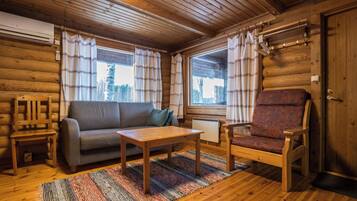 Interno di una baita di legno con divano, poltrona, tavolo e vista all'esterno tramite ampie finestre.