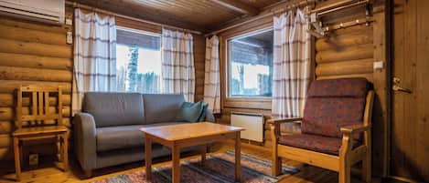 A log cabin interior with a sofa, armchair, table, and a view of the outside through large windows.
