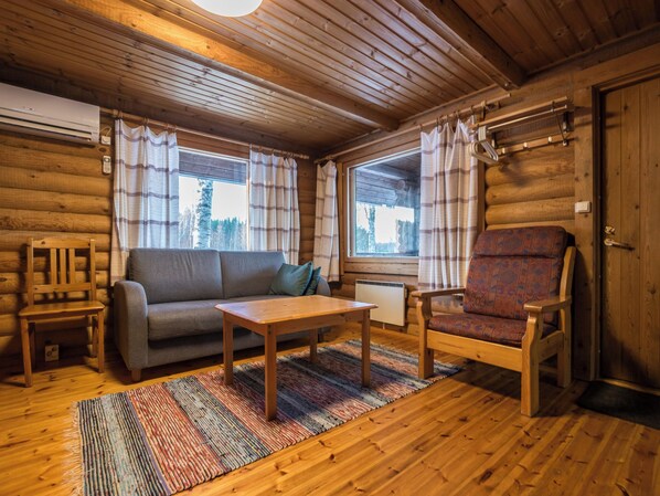 A log cabin interior with a sofa, armchair, table, and a view of the outside through large windows.