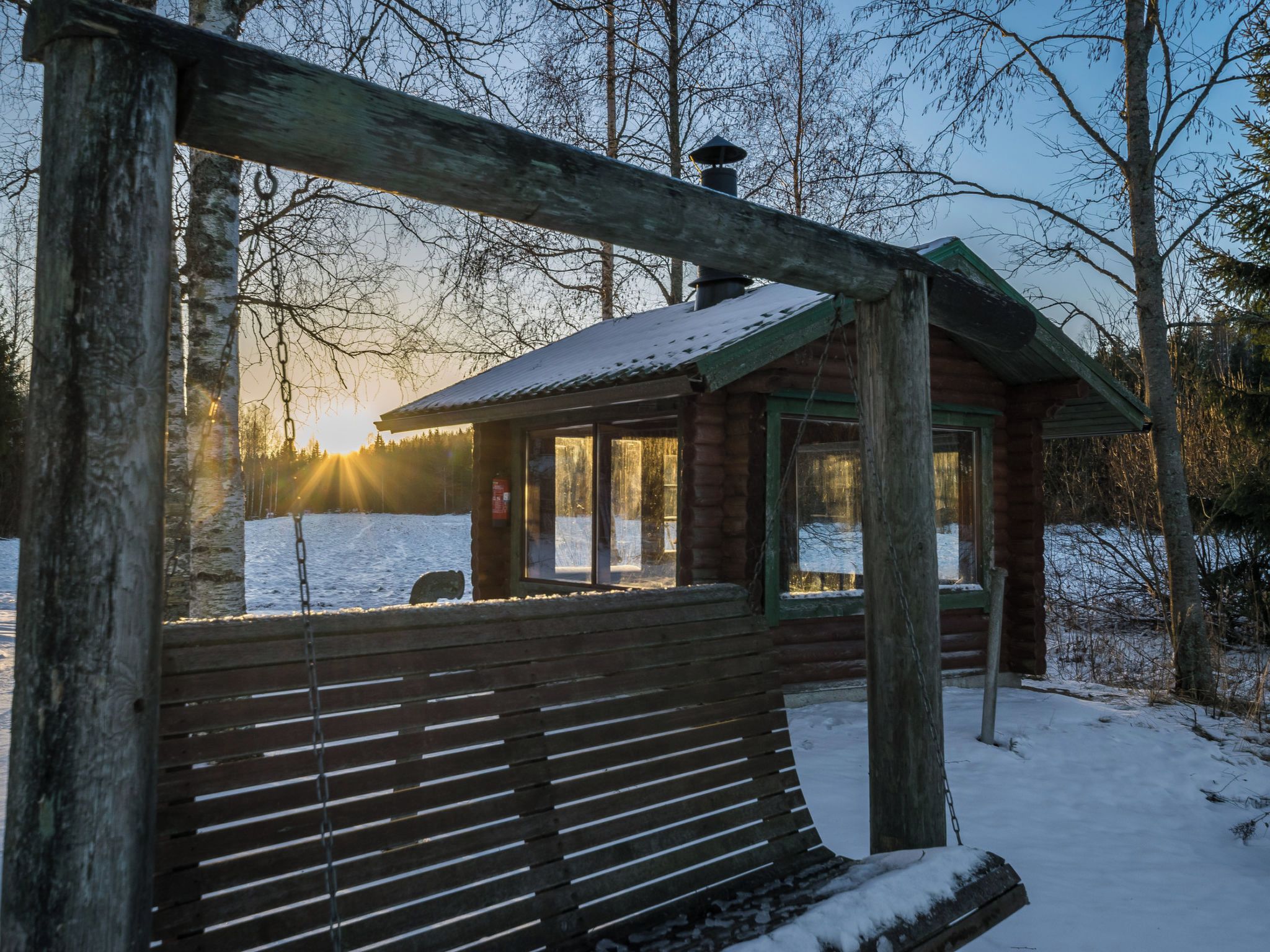 A wooden cabin with a bench in front, surrounded by snow-covered ground and trees.