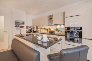 A modern kitchen with a central island, a refrigerator, and a dining area with two chairs. - Ferienwohnung Lieblingsort - Modern beach apartment with balcony & 2 bathrooms (Loddin)