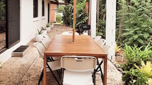 A wooden table with white chairs and a large umbrella in a garden area with plants and a white building in the background.