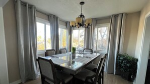 Een eetkamer met een rechthoekige tafel, zes stoelen, een kroonluchter en grote ramen met gordijnen.