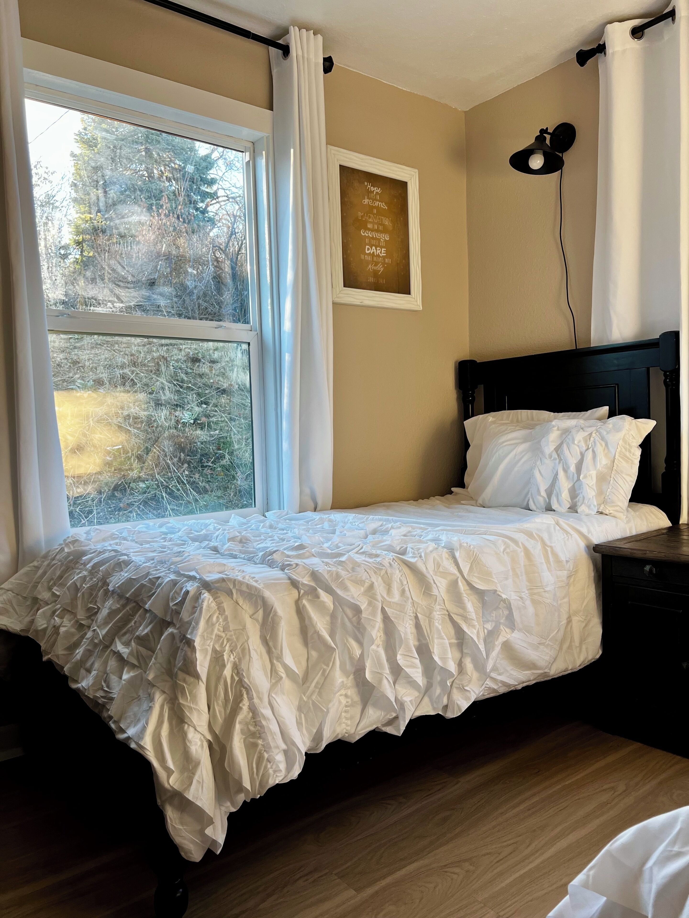 Un dormitorio con cama, mesita de noche, ventana con cortinas, lámpara de pared y una cita motivacional en la pared.