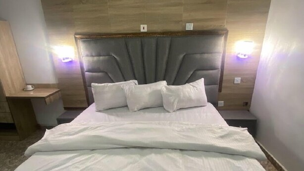 A neatly made bed with a grey upholstered headboard, two bedside lamps, and a wooden panel wall. - DE EMERITUS COURTYARD HOTEL (Abuja)