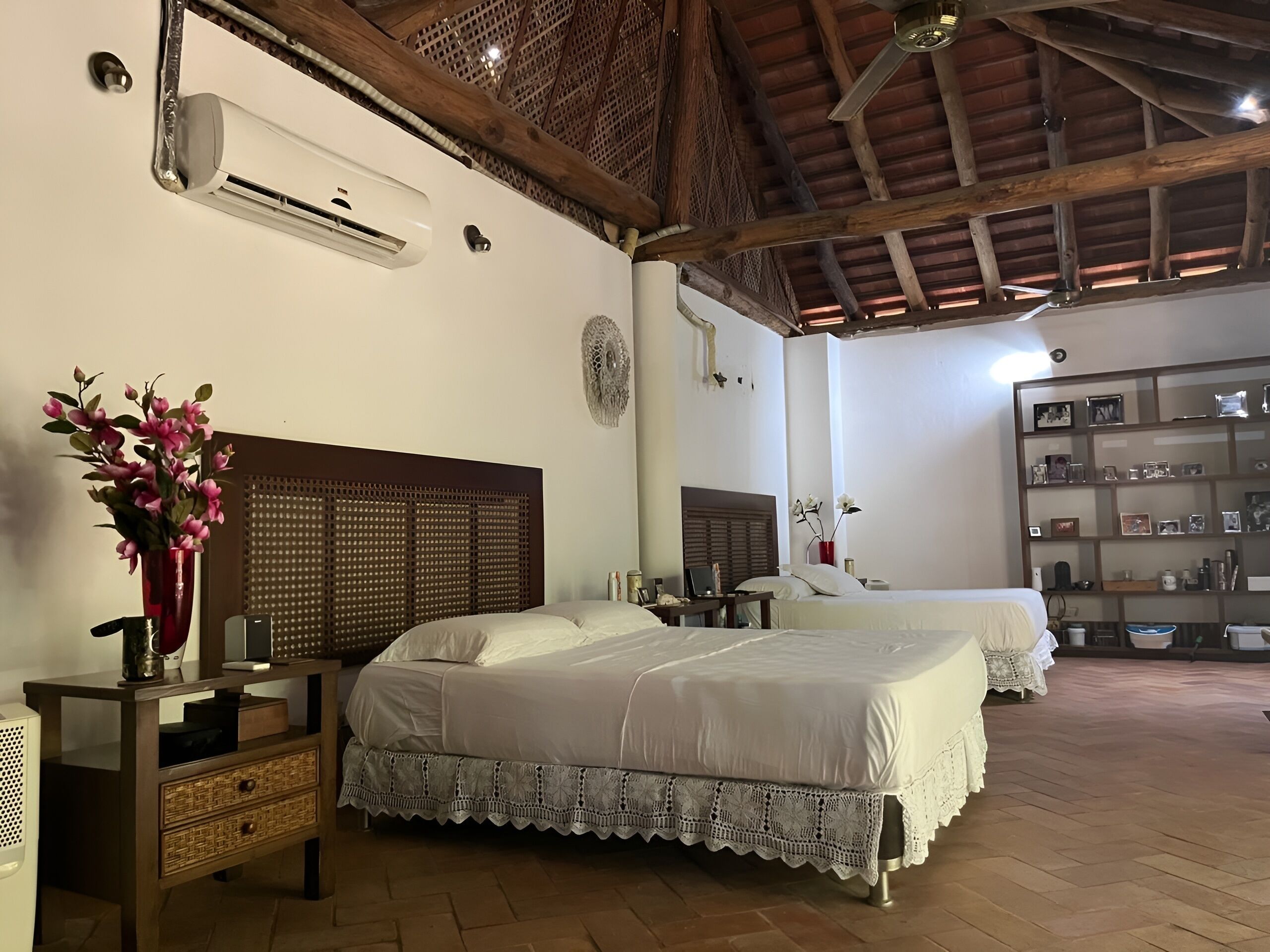 Un dormitorio con dos camas, un cabecero de madera, una mesita de noche con un florero y una estantería con diversos objetos.
