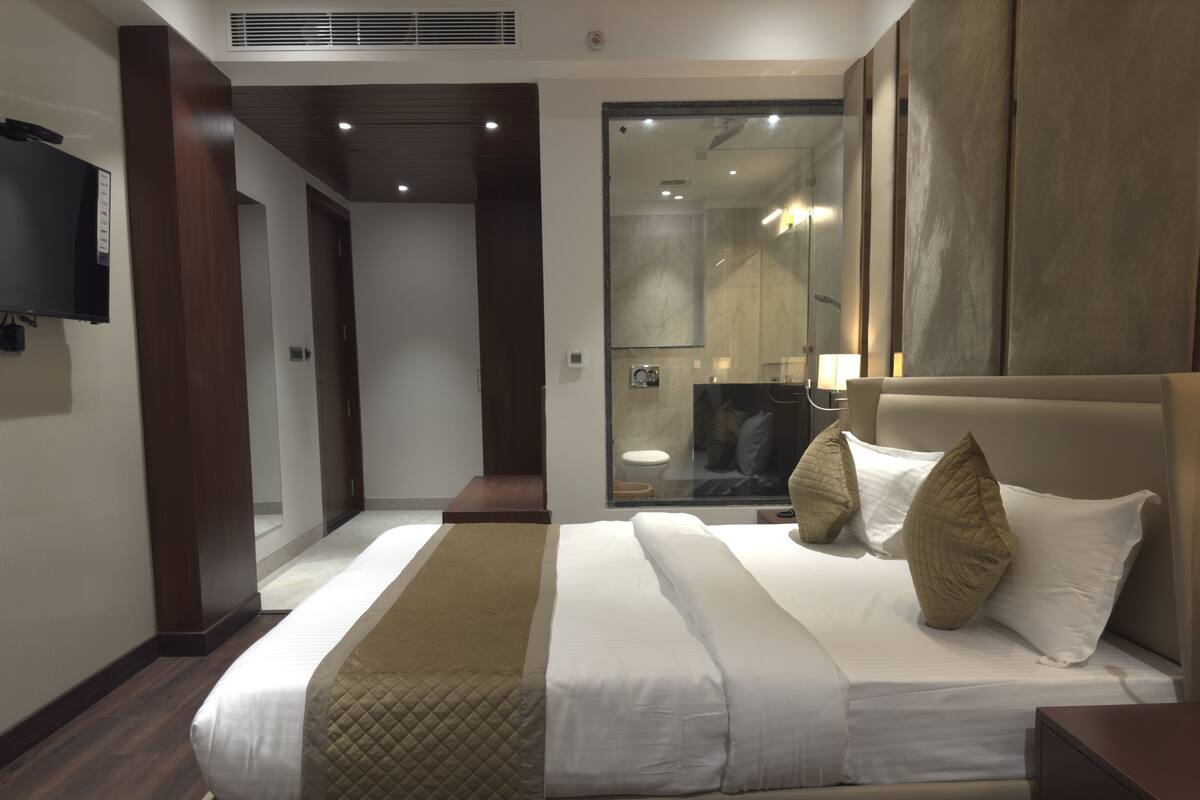 A modern hotel room with a large bed, a television mounted on the wall, and a bathroom visible through a glass partition.