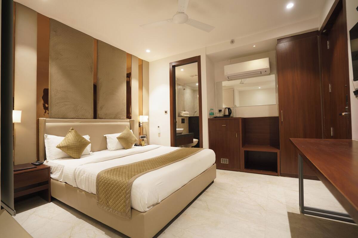 A modern hotel room with a large bed, a desk, a wardrobe, and a bathroom visible through an open door.