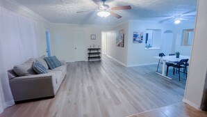 A spacious living room with a wooden floor, a ceiling fan, a dining area with a table and chairs, and a door leading to another room. - Charming 3-bedroom house in delightful Jacksonville with AC, WiFi (Jacksonville)