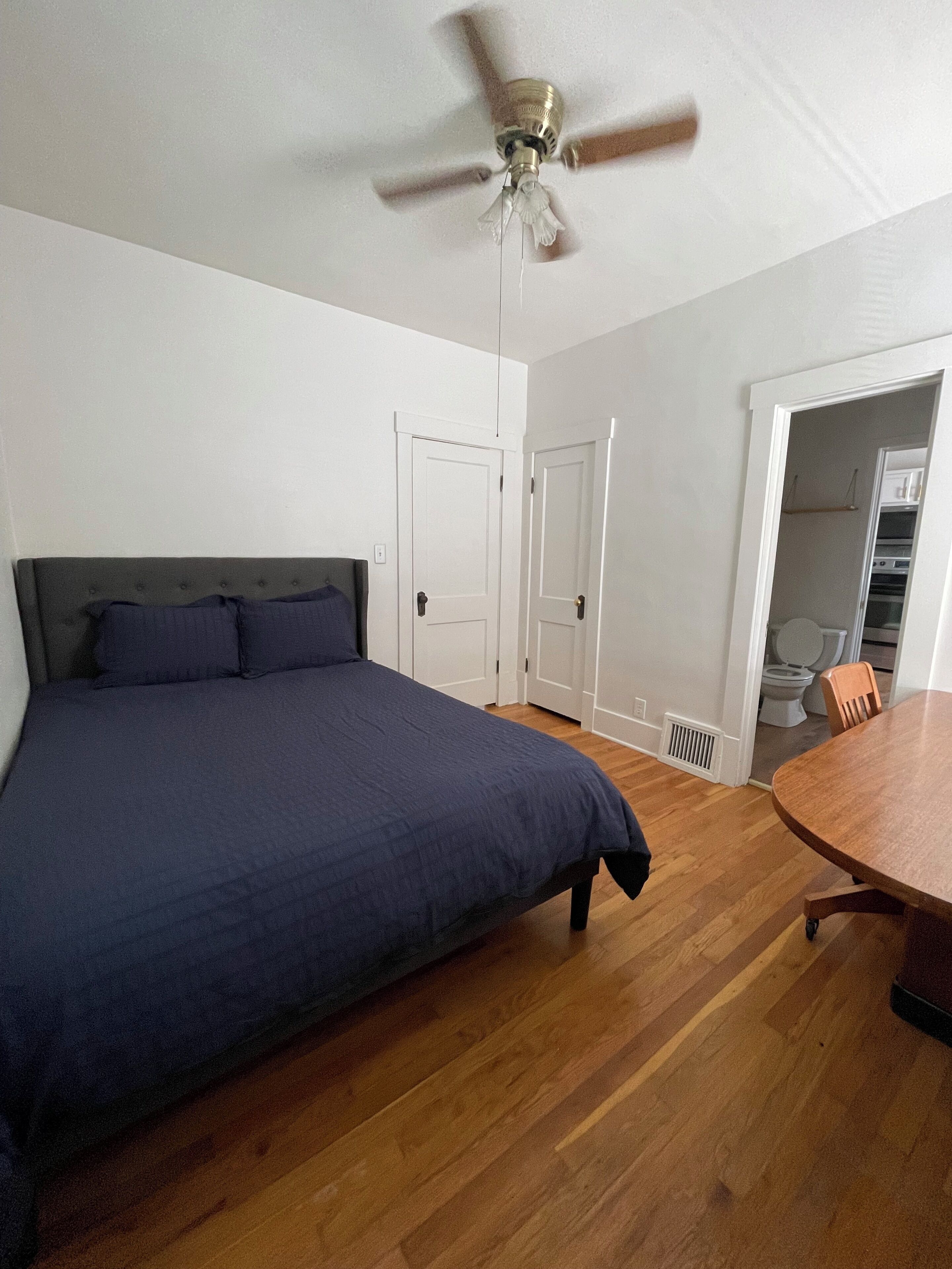 A bedroom with a bed, a ceiling fan, a wooden dining table, and a bathroom visible through an open door.