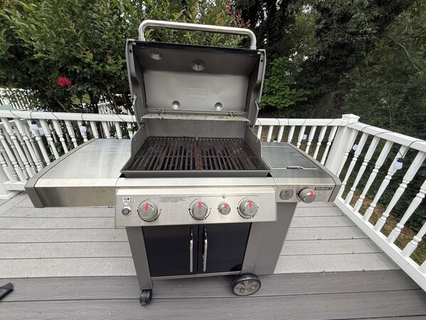 A portable gas grill with four burners and a side burner, placed on a wooden deck with white railings.