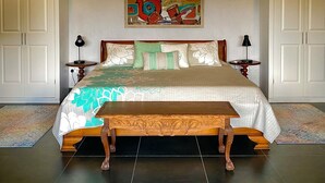 Una cama grande con colcha floral, dos mesitas de noche con lámparas, un banco y un cuadro abstracto colorido sobre la cama.