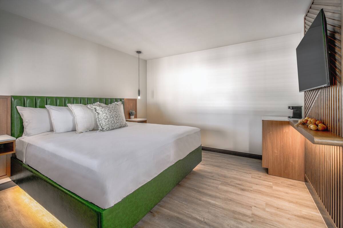 Une chambre d’hôtel moderne avec un grand lit, une table de chevet en bois, une petite table avec des fruits et un téléviseur à écran plat fixé au mur.