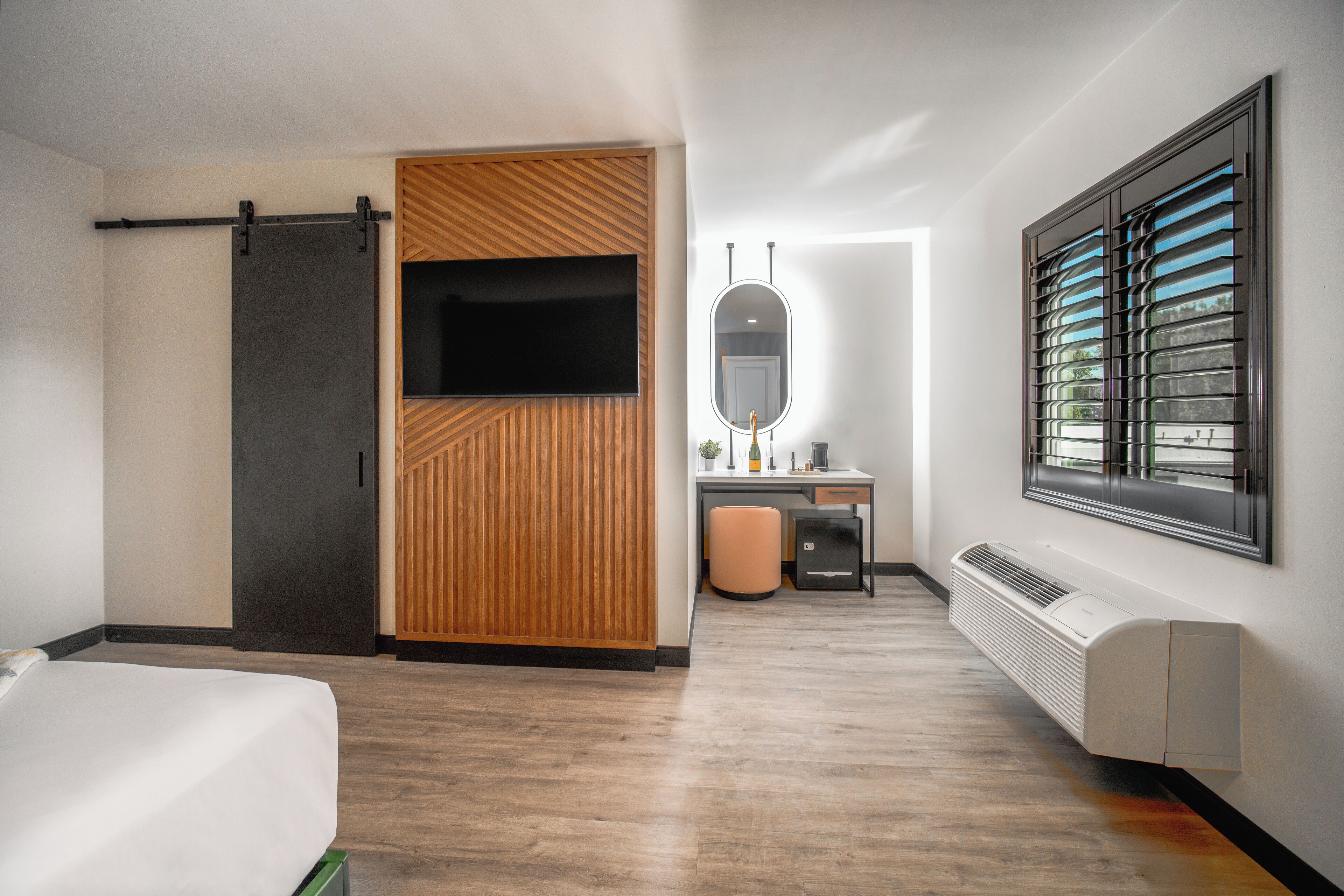 A modern hotel room with a bed, a desk with a chair, a mirror, a sliding door, and a window with blinds.