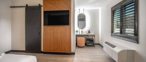 A modern hotel room with a bed, a desk with a chair, a mirror, a sliding door, and a window with blinds.