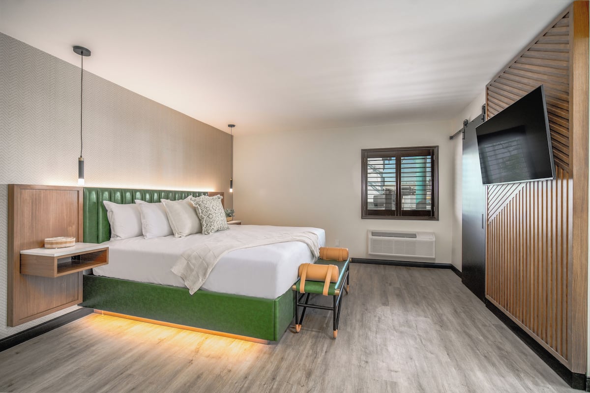 Une chambre d’hôtel moderne avec un lit recouvert d’un tissu vert, une table de chevet en bois, un banc, un téléviseur à écran plat et une fenêtre équipée de stores.