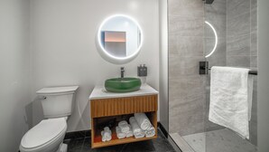 A modern bathroom with a toilet, a green sink, a wooden vanity, a round mirror with lights, and a glass shower enclosure.
