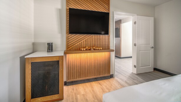 A hotel room with a wooden accent wall, a flat-screen TV mounted above a wooden console, a small coffee maker, and a bed with white bedding.