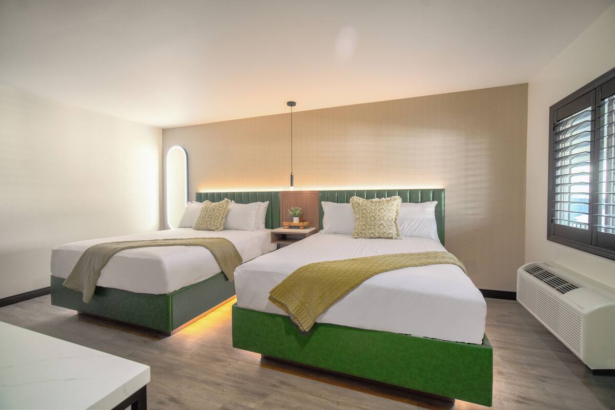 Une chambre d’hôtel avec deux lits, une table de chevet, une lampe et une fenêtre avec des stores.