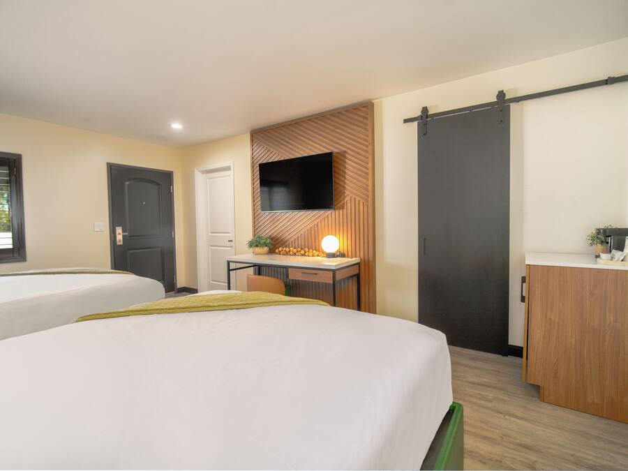 Quarto de hotel com duas camas, uma escrivaninha com televisão, uma porta de correr e uma kitchenette pequena.