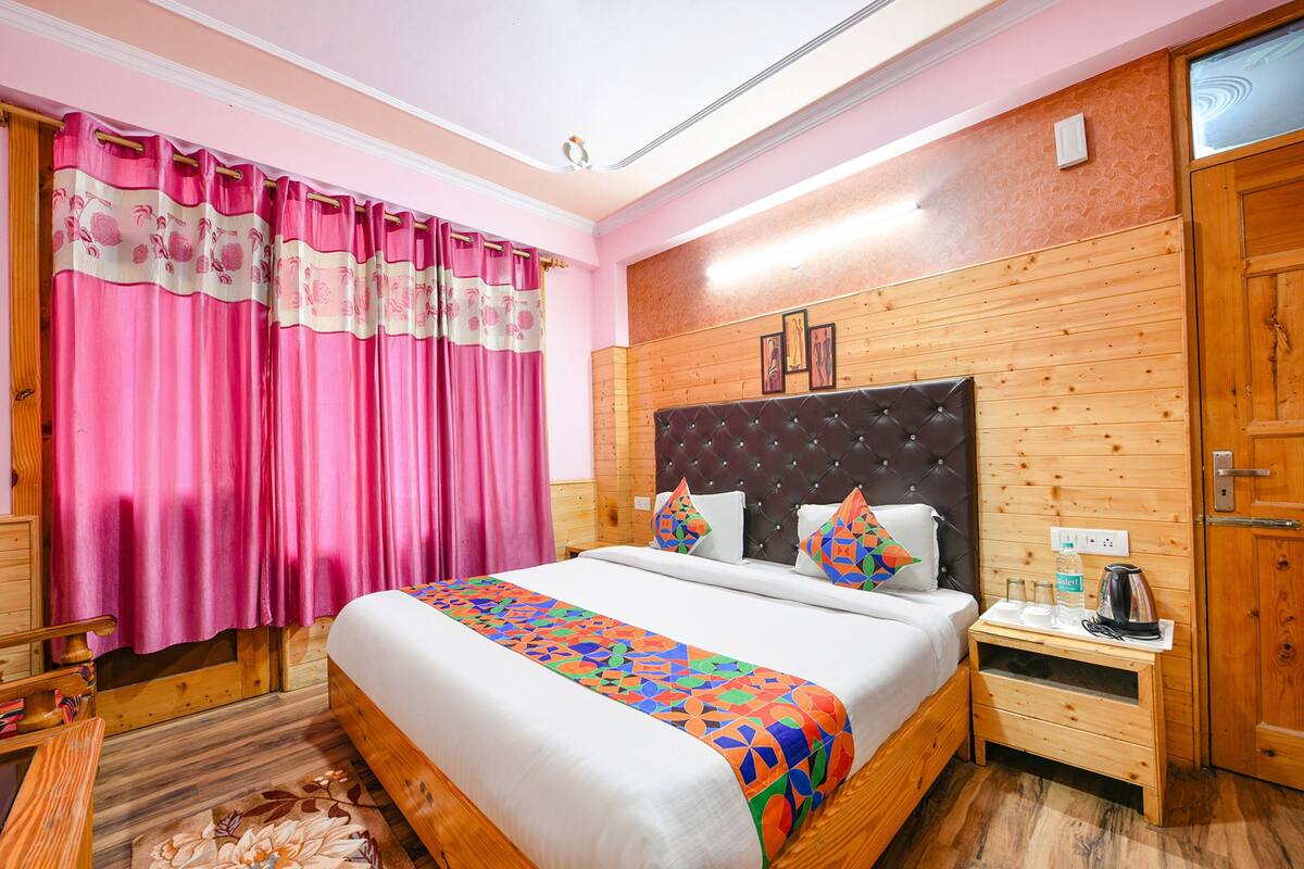 A bedroom with a large bed, wooden headboard, pink curtains, and a wooden nightstand with a kettle.