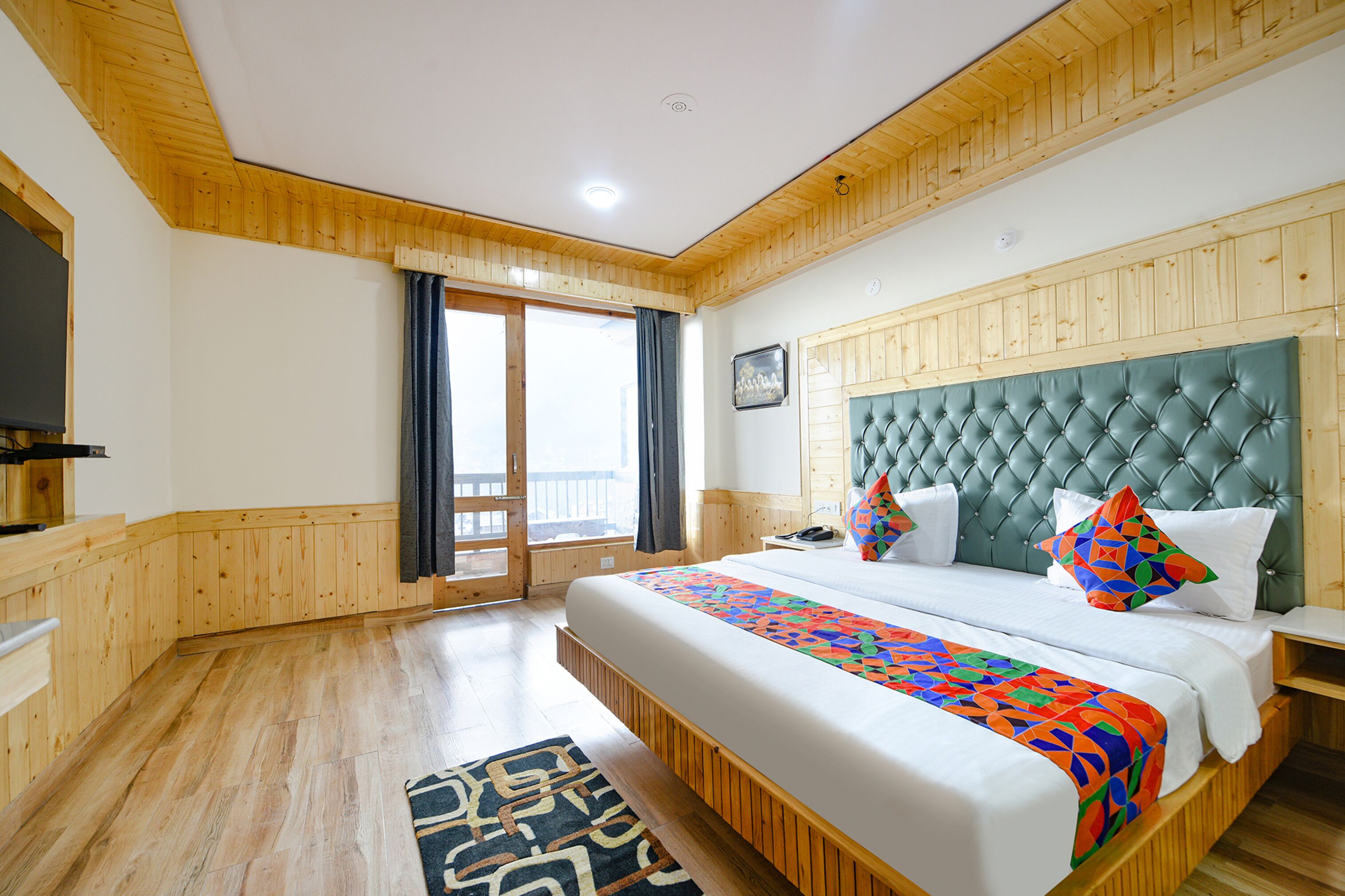 A hotel room with a large bed, wooden paneling, a view through a sliding door, and a patterned rug.